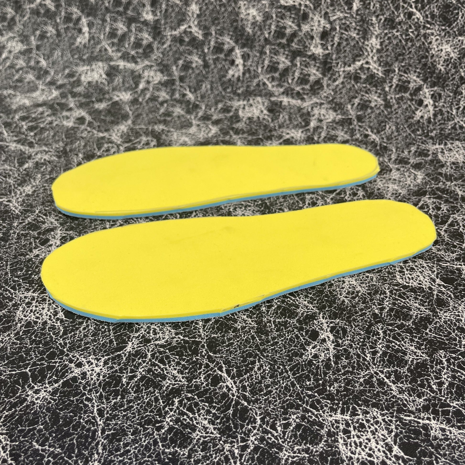 two cut out insoles upside down on a sheet of Silver AG 2.0 footbed Nylon fabric. Insole is the 1/4" sheet that has 3 layers, the black thin footbed layer, the 1/8"BounceBlu Layer and the 1/8" VeloShox Layer.