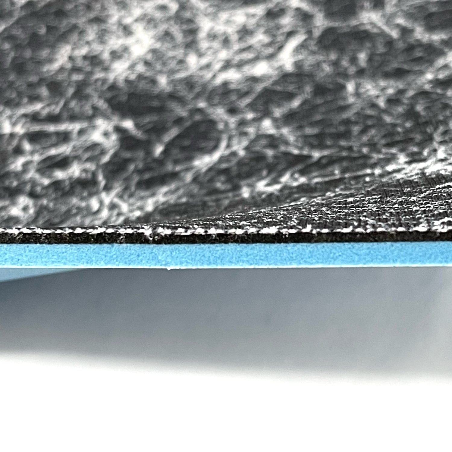 Side profile of insole sheet combination. You can see the silver infused black with silver pattern footbed on top and underneath that a thin 1/16" Black layer of FoamFuzion, then a 1/8" Blue layer of bounceBlue on a white background.